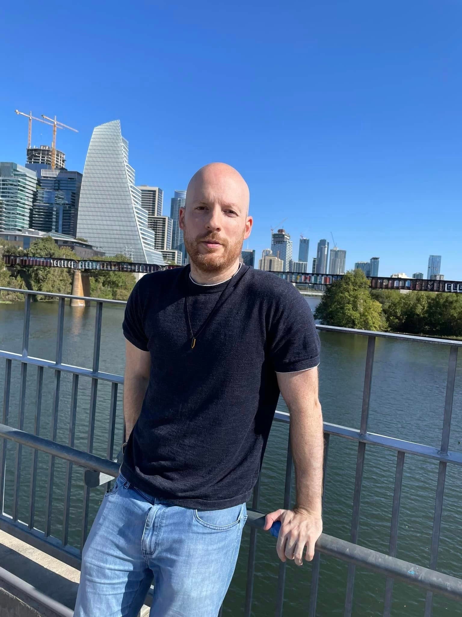 A bald man with a short beard stands by a metal railing along a river, wearing a dark short-sleeve t-shirt and light blue jeans, with a small pendant necklace. In the background is a modern city skyline with tall buildings and a clear blue sky.
