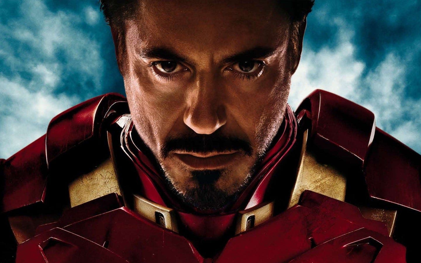 Close-up of Tony Stark in red and gold Iron Man armor, looking downward with a stern, focused expression. The blue, cloudy sky forms the dramatic background.